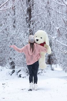 Woman standing in the snow covered pine forest in winter with snow falling all around her.  She is wearing a pink crochet knit jumper and holding a large oversized soft toy