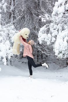 Happy go lucky woman frolicking in snow lifting playfully a large soft toy above her head in a snow covered forest of pines in winter