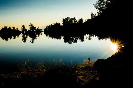 Sunset silhouette of trees and lake in natural enviornment, blue sky, reflections of surrounding landscape.