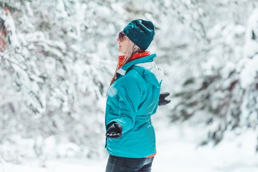 Woman wearing winter jacket and beanie standing out in falling snow  among a forest of pine trees