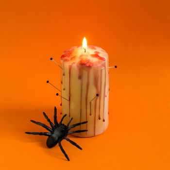 Homemade decor for Halloween. Candle pierced with needles with red brooks and drops like blood and spider on orange background. Minimal style. Original design