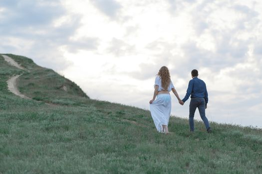 A couple in love, a pregnant wife in a white outfit are walking in a hilly field. Overcast weather,