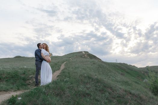 A couple in love, a pregnant wife in a white outfit are walking in a hilly field. Overcast weather,