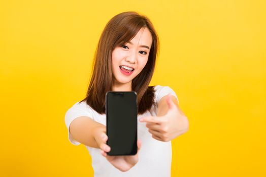 Asian happy portrait beautiful cute young woman smile standing wear t-shirt making finger pointing on smartphone blank screen looking to phone isolated, studio shot yellow background with copy space