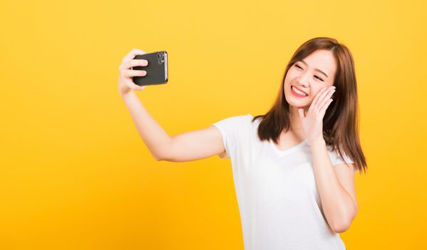 Asian happy portrait beautiful cute young woman teen smiling standing making selfie photo, video call on smartphone raise hand to say hello isolated, studio shot yellow background with copy space