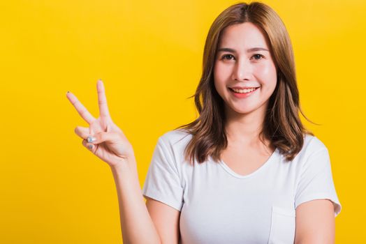 Asian Thai happy portrait beautiful cute young woman smile standing wear t-shirt showing finger making v-sign symbol near eye looking to camera, isolated studio shot yellow background with copy space