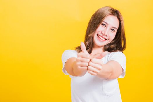 Portrait Asian Thai beautiful happy young woman smiling wear white t-shirt standing successful woman giving two thumbs up gesture sign in studio shot, isolated on yellow background with copy space