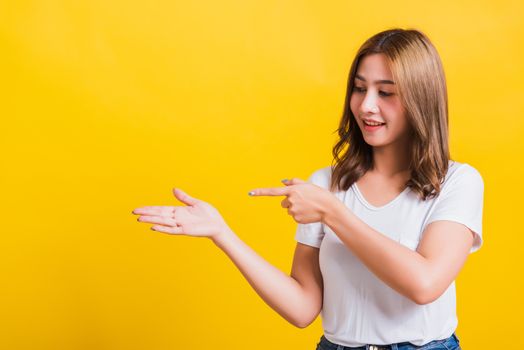 Asian Thai happy portrait beautiful young woman standing to hold something on palm away side and point the finger to it looking to side, studio shot isolated on yellow background with copy space