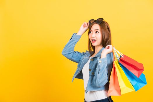 Asian happy portrait beautiful cute young woman teen smiling standing with sunglasses excited holding shopping bags multi color looking camera isolated, studio shot yellow background with copy space