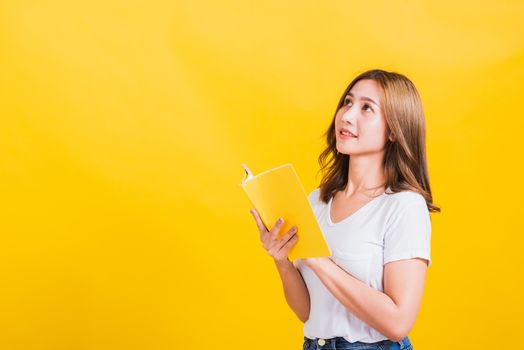 Portrait Asian Thai beautiful happy young lifestyle woman stands holding yellow book or diary she show thumb up finger and looking up away, studio shot isolated on yellow background, with copy space