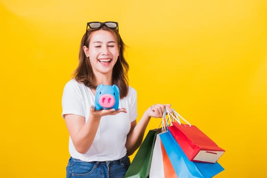 Portrait Asian Thai beautiful young woman happy hold colorful shopping bags and Piggybank Saving money, studio shot isolated on yellow background, with copy space