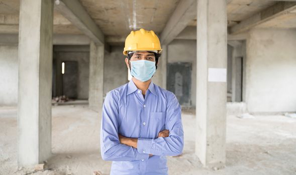 Concept of back to work, opening of construction sites after covid-19 pandemic - portrait of confident construction worker in a construction helmet arms crossed with medical mask at site.