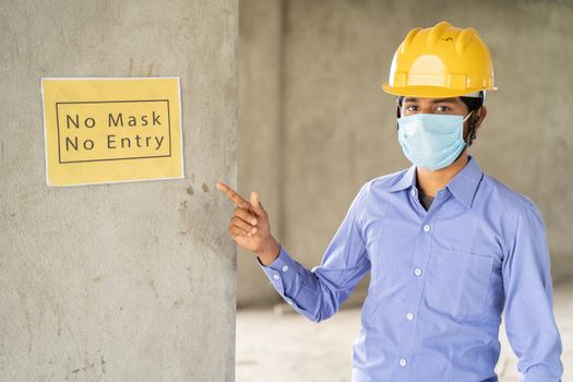 worker pointing No Mask No Entry signage notice board on wall at working construction site to protect from coronavirus or covid-19 at workplaces - concept of health and labor safety during pandemic.