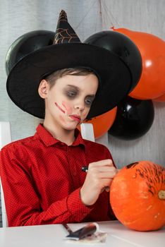 Happy teen boy in costume preparing for the Halloween celebration drawing a pumpkin. Halloween carnival or masquerade concept