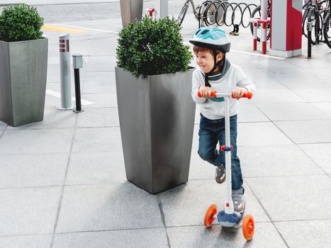 Toddler in helmet is riding scooter on parking lot with parked bicycles. Urban vehicle for active children. Leisure activity for boys and girls.