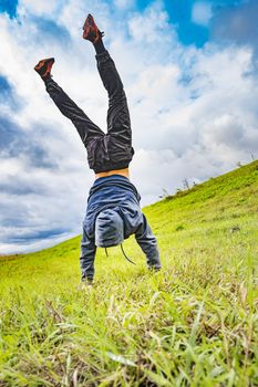 a man, a boy, a teenager stands on his hands upside down on the grass on a mountainside against the background of the sky and clouds