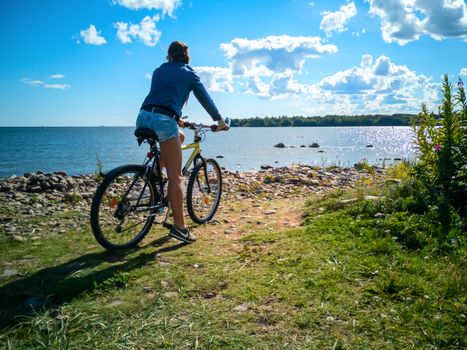 Young Woman Cyclist Riding Road Bike with blue sky and clouds on background. Adventure, Healthy Lifestyle, Sport concept. Girl with bicycle on the coast of the baltic sea. Sky dreamy travel photo. Copy space.