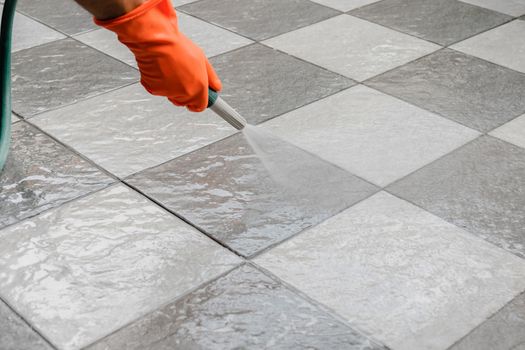 Hand of man wearing orange rubber gloves is use a hose to clean the tile floor.