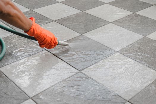 Hand of man wearing orange rubber gloves is use a hose to clean the tile floor.