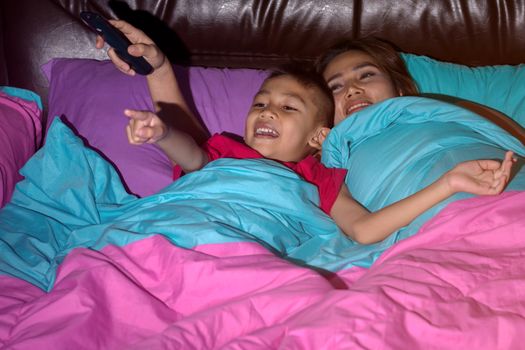 Mother and son sleeps on the bed and holds the TV remote and smiles happily, concepts of technology and entertainment.