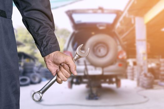 Car repairman wearing a dark blue uniform standing and holding a wrench that is an essential tool for a mechanic and has a backdrop as a car repair center.