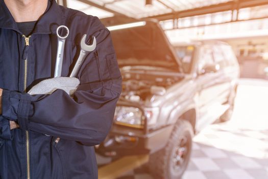 Car repairman wearing a dark blue uniform standing and holding a wrench that is an essential tool for a mechanic and has a backdrop as a car repair center.