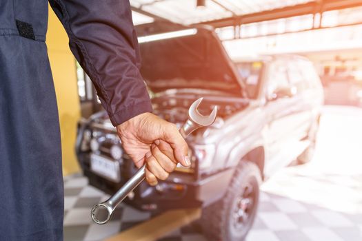 Car repairman wearing a dark blue uniform standing and holding a wrench that is an essential tool for a mechanic and has a backdrop as a car repair center.