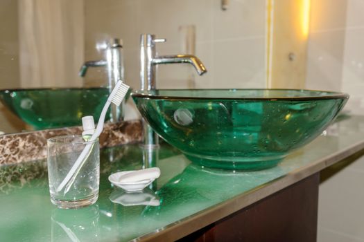 Toothbrush and toothpaste in a glass placed in front of the mirror near the basin in the bathroom.