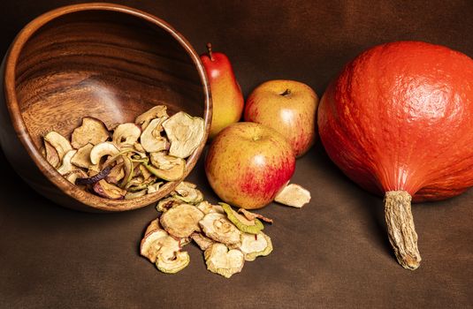 Still life of fresh red kuri squash, poppy heads and a pile of dried apples in a wooden bowl together with two whole apples and one pear. Still life concept of fall season harvest and homemade fruit processing ona brown background