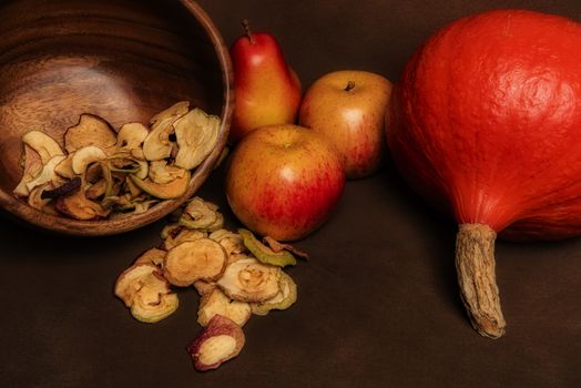 Still life of fresh red kuri squash, poppy heads and a pile of dried apples in a wooden bowl together with two whole apples and one pear. Still life concept of fall season harvest and homemade fruit processing ona brown background