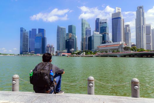 Asian men carrying backpack are sitting by the river and looking to the building at the center of business in the big city.