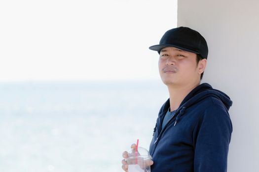 Asian man wearing a blue long sleeve shirt and wearing a hat holding a plastic glass of water by the ocean