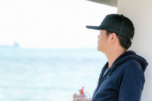 Asian man wearing a blue long sleeve shirt and wearing a hat holding a plastic glass of water by the ocean