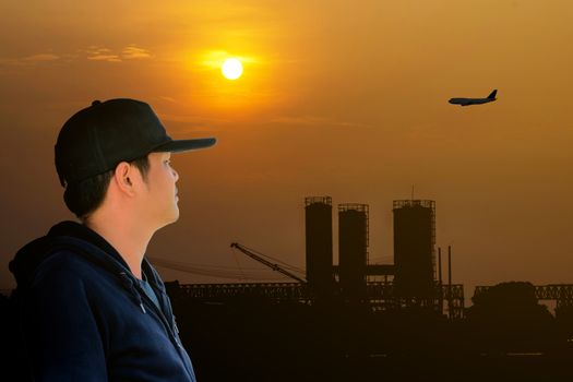 Asian man wearing a blue long sleeve shirt and wearing a hat looking at the airplane flying over the factory on the red sky with beautiful sunlight.