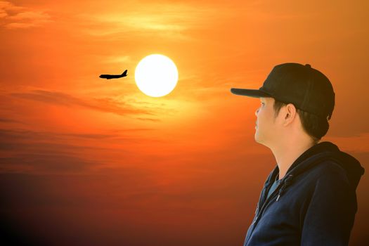 Asian man wearing a blue long sleeve shirt and wearing a hat looking at the airplane on the red sky with beautiful sunlight.