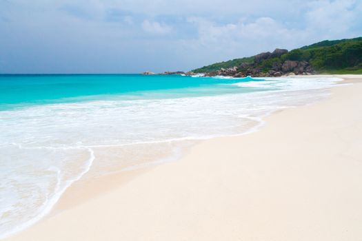 White sand beach with some waves and blue sky in the far a hill with green plants