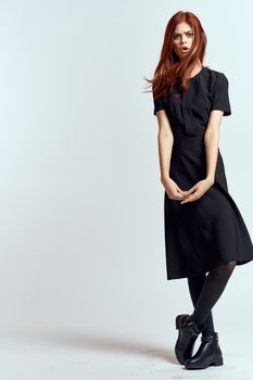 A woman in a black dress on a light background and pantyhose shoes red hair and pose in full growth. High quality photo