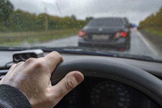 the driver's hand on the steering wheel of the car against the background of the car in front, which is waiting for the signal to avoid an obstacle due to road repairs in rainy cloudy weather in the autumn time period