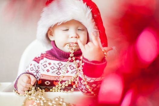 A little baby girl in a New Year's hat of Santa Claus examines and plays with New Year's decorations. Merry Christmas and Happy New Year greetings.