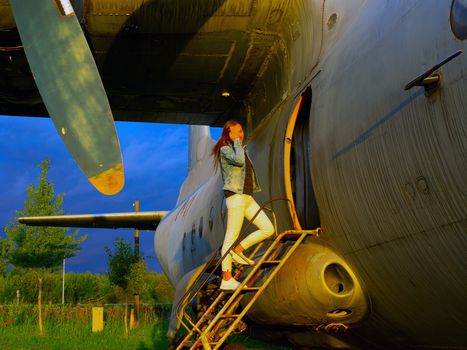 Yo ungwoman standing at plane ladder going to board, outdoors, airport. Old Soviet military airplane, sunset time. Close up of a Abandoned Historic AircraftAN-12. Close up of propeller engine.