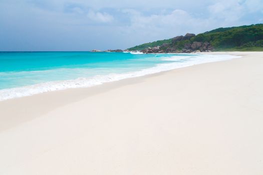 White sand beach with some waves and blue sky in the far a hill with green plants