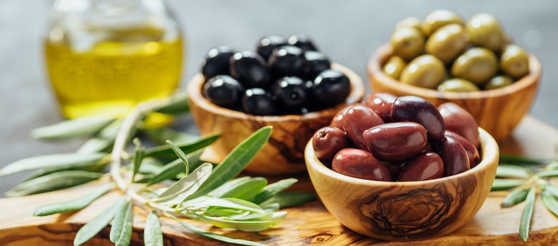 Set of green, red and black olives and olive oil on gray background. Different types of olives in olive wooden bowls and olive oil on wooden cutting board and olive leaves.Copy space.Horizontal banner