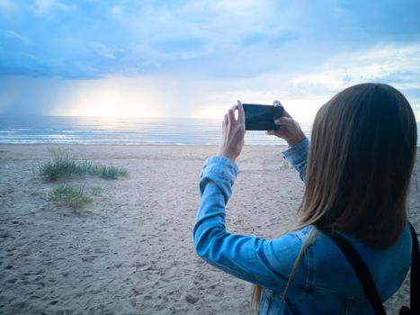 beautiful alone girl on the beach. Girl looking at stormy sea and taking a picture with smarphone. The spectacular Storm with rain Is Coming in Estonia. Baltic sea