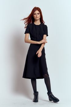 A woman in a black dress on a light background and pantyhose shoes red hair and pose in full growth. High quality photo