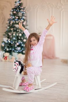 A beautiful little girl in pink pajamas rejoices in a wooden rocking horse, a gift from Santa for Christmas.