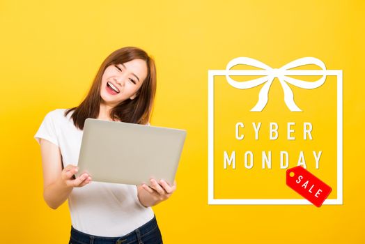 Asian happy portrait beautiful cute young woman teen smile standing hold laptop computer and excited celebrating success with Cyber Monday in the gift box on space, studio shot yellow background