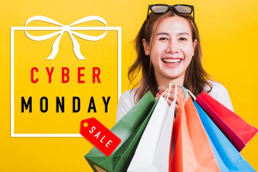 Asian happy portrait beautiful young woman smiling stand with sunglasses excited holding shopping bags multi color looking camera with Cyber Monday text in gift box on side isolated yellow background