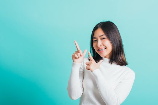 Portrait of Asian teen beautiful young woman smile have dental braces on teeth laughing point finger side away blank copy space, studio shot isolated on blue background, medicine and dentistry concept