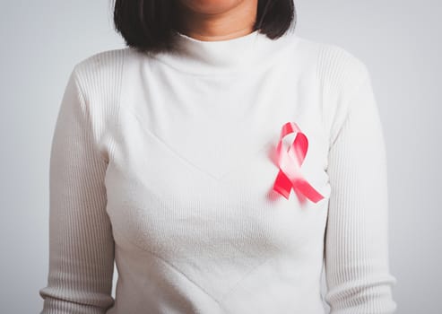 Breast cancer awareness healthcare and medicine concept. Close up Asian woman wear white shirt standing with pink breast cancer awareness ribbon pin on chest, studio shot isolated on white background