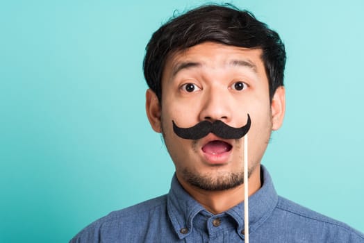 Portrait Asian happy handsome man posing he holding a funny mustache card or vintage fake moustaches on his mouth, studio shot isolated on blue background, Fathers day, November day concept